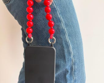 Pode incluir: Uma capa de telefone preta com uma alça de contas vermelhas. A alça é feita de contas vermelhas grandes, redondas e brilhantes. A capa do telefone tem um módulo de câmera com três lentes.