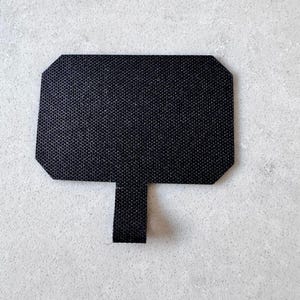 May include: A black rectangular patch with a small tab extending from the bottom. The patch has a textured surface and a geometric shape with beveled corners. The background is a light grey, speckled surface.