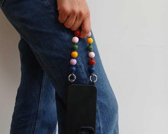 May include: Black phone case with a colorful beaded strap. The strap features a variety of round beads in red, pink, yellow, green, purple, and blue. The phone case is held by a person wearing blue jeans.
