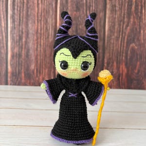 May include: A crocheted amigurumi doll of Maleficent from Disney's Sleeping Beauty. The doll is green, black, and purple with a yellow staff.