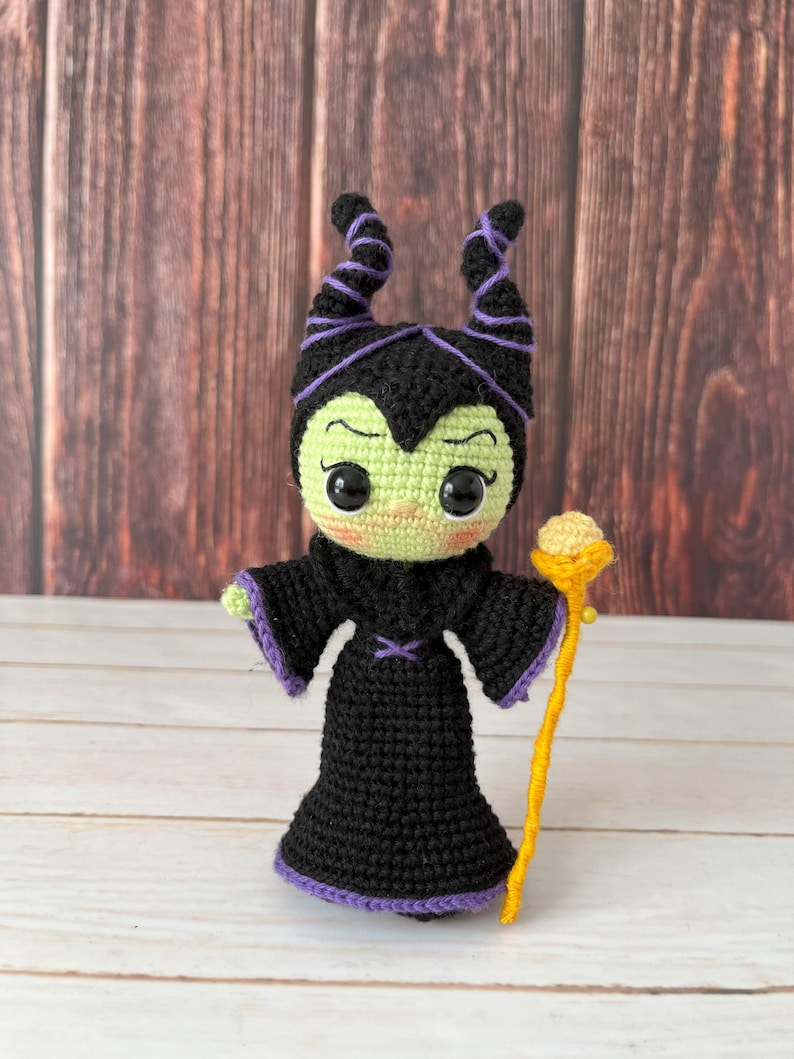 May include: A crocheted amigurumi doll of Maleficent from Disney's Sleeping Beauty. The doll is green, black, and purple with a yellow staff.