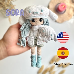 Puede incluir: Una muñeca de crochet llamada Sora que lleva un gorro blanco con ribete azul, un suéter blanco y zapatos azules. La muñeca sostiene un conejito blanco de crochet. La muñeca está sentada en una mano.