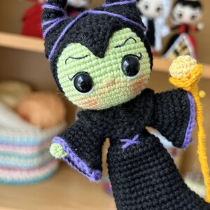 May include: A green and black crocheted doll with purple horns and a yellow staff. The doll has large black eyes and a mischievous expression.