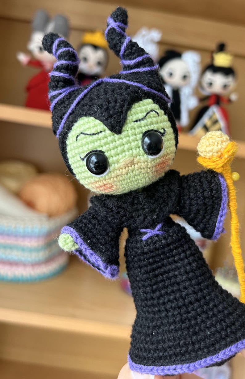May include: A green and black crocheted doll with purple horns and a yellow staff. The doll has large black eyes and a mischievous expression.