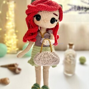 May include: A handmade crochet doll with red hair, green accents, and a light green dress. The doll has a small purse and green sandals. The doll is a whimsical, handcrafted item.