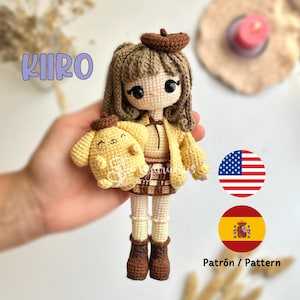 Könnte beinhalten: Häkel-Amigurumi-Puppe mit braunen Haaren, die eine gelbe Jacke, einen braunen Rock und braune Stiefel trägt. Die Puppe hält einen gelben gehäkelten Welpen. Die Puppe ist mit "KIRO" beschriftet.
