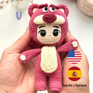 May include: Crocheted amigurumi toy of a character wearing a pink bear costume. The toy has large eyes, a small nose, and a yellow belly. The toy is being held in a hand with the words "Patrón / Pattern" and flags of the United States and Spain in the background.
