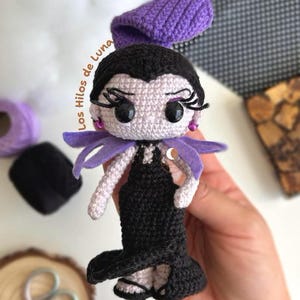 K&ouml;nnte beinhalten: Eine geh&auml;kelte Puppe mit schwarzen Haaren, einem lila Hut und einem schwarzen Kleid. Die Puppe hat gro&szlig;e Augen und einen schelmischen Ausdruck. Die Puppe h&auml;lt ein lila Band in der Hand.