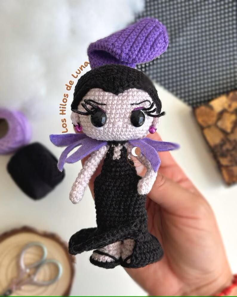 K&ouml;nnte beinhalten: Eine geh&auml;kelte Puppe mit schwarzen Haaren, einem lila Hut und einem schwarzen Kleid. Die Puppe hat gro&szlig;e Augen und einen schelmischen Ausdruck. Die Puppe h&auml;lt ein lila Band in der Hand.