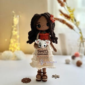 May include: Crocheted doll with brown hair, wearing a white dress with a brown and orange pattern, holding a small white crocheted animal. The doll is standing on a white surface with a red flower.
