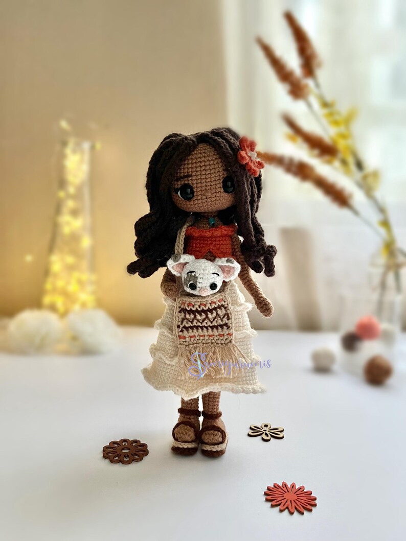 May include: Crocheted doll with brown hair, wearing a white dress with a brown and orange pattern, holding a small white crocheted animal. The doll is standing on a white surface with a red flower.