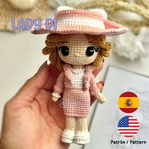 May include: A crocheted doll wearing a pink and white striped hat, a pink jacket, and a pink skirt. The doll has brown hair and large brown eyes. The doll is holding a small white bag. The doll is standing on a hand. The image also includes the text "LADY DI" and the flags of Spain and the United States.