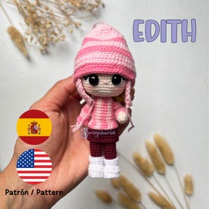 Puede incluir: Un gorro de punto de rayas rosa y blanco con un jersey de punto rosa y pantalones burdeos. La muñeca de ganchillo tiene ojos negros y botas blancas. La muñeca está etiquetada como "Edith".
