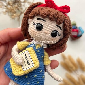 May include: Crocheted doll of a girl wearing a blue dress with yellow sleeves and a red bow. The doll is holding a yellow book labeled "MOBY DICK" by Herman Melville. The doll has brown hair and white socks.