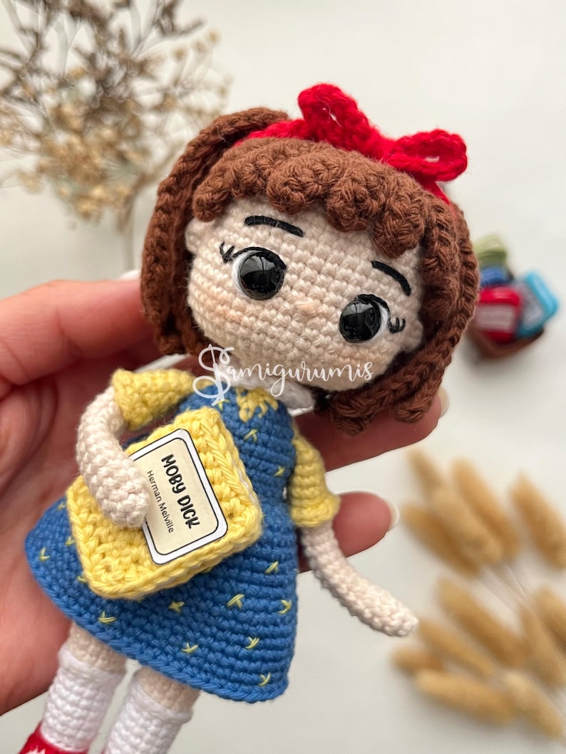 May include: Crocheted doll of a girl wearing a blue dress with yellow sleeves and a red bow. The doll is holding a yellow book labeled "MOBY DICK" by Herman Melville. The doll has brown hair and white socks.