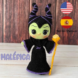 以下が含まれることがあります： 紫色の角と緑色の肌、黒いドレスを着た、クロシェで編まれたマレフィセント人形。人形は黄色の杖を持っています。画像には「MALÉFICA」というテキストが表示されています。