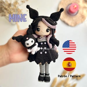 Puede incluir: Una muñeca de crochet negra y blanca con pelo rosa y un vestido negro. La muñeca sostiene un pequeño animal de crochet negro y blanco. La muñeca está etiquetada como "MINE".