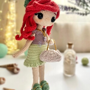 May include: A handmade crochet doll with red hair, wearing a green skirt, purple top, and green shoes. The doll has a small purse and a seashell necklace. The doll is approximately 8 inches tall.