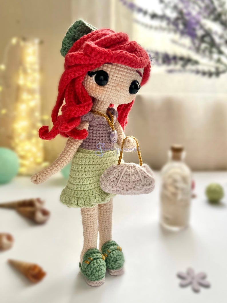May include: A handmade crochet doll with red hair, wearing a green skirt, purple top, and green shoes. The doll has a small purse and a seashell necklace. The doll is approximately 8 inches tall.