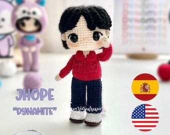 May include: Crocheted doll of J-Hope from the group BTS, wearing a red jacket, dark blue pants, and white shoes. The doll has black hair and large eyes. The text "JHOPE" and "Dynamite" are visible. The image also includes a Spanish and American flag.