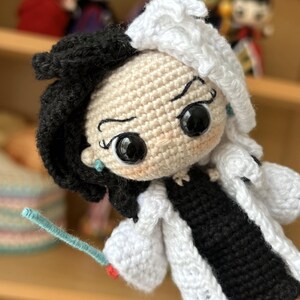 May include: Crocheted doll with black and white hair, wearing a white coat and black dress. The doll has large black eyes and a mischievous expression. The doll is holding a blue wand.