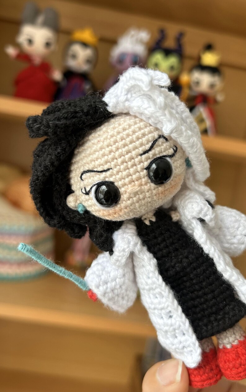 May include: Crocheted doll with black and white hair, wearing a white coat and black dress. The doll has large black eyes and a mischievous expression. The doll is holding a blue wand.