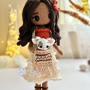 May include: Crocheted doll with brown hair, wearing a white dress with a brown and orange patterned vest, holding a small white crocheted animal.