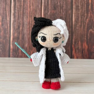 May include: A crocheted doll with black and white hair, wearing a white coat and a black dress. The doll is holding a blue wand and has red shoes.
