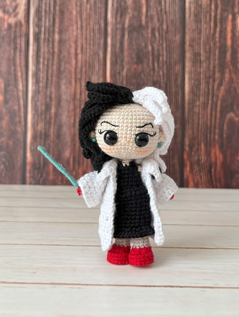 May include: A crocheted doll with black and white hair, wearing a white coat and a black dress. The doll is holding a blue wand and has red shoes.