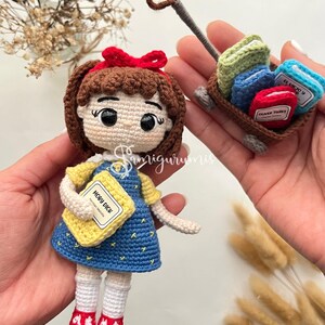 May include: A handmade crochet doll of a girl in a blue dress, holding a yellow book labeled "Moby Dick." A small wagon carries miniature books labeled "Oliver Twist" and "El Viejo y El Mar." The doll has a red bow and red shoes.