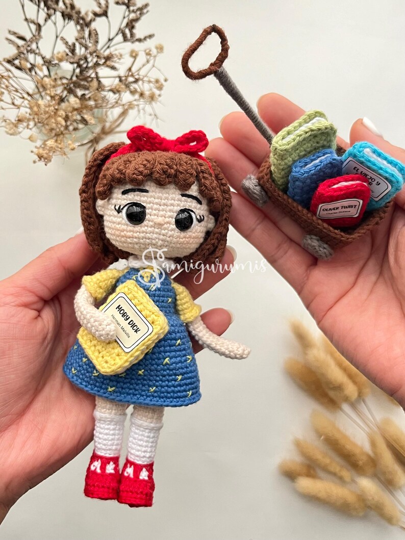 May include: A handmade crochet doll of a girl in a blue dress, holding a yellow book labeled "Moby Dick." A small wagon carries miniature books labeled "Oliver Twist" and "El Viejo y El Mar." The doll has a red bow and red shoes.