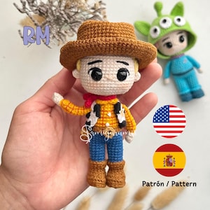 May include: Crocheted amigurumi doll dressed as Woody from Toy Story. The doll has a brown cowboy hat, a yellow and red plaid shirt, a brown vest with a sheriff's star, blue jeans, and brown boots. The doll is holding its right arm out, as if waving. The doll is standing on a hand, and there are American and Spanish flags in the background.
