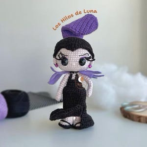 K&ouml;nnte beinhalten: H&auml;kelpuppe Amigurumi mit schwarzen Haaren, einem lila Hut und einem schwarzen Kleid. Die Puppe hat gro&szlig;e Augen und einen s&uuml;&szlig;en Ausdruck. Die Puppe steht auf einer wei&szlig;en Oberfl&auml;che. Der Text "Los Hilos de Luna" ist in Orange &uuml;ber der Puppe geschrieben.