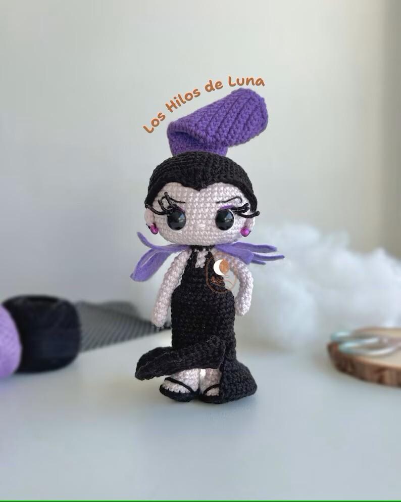 K&ouml;nnte beinhalten: H&auml;kelpuppe Amigurumi mit schwarzen Haaren, einem lila Hut und einem schwarzen Kleid. Die Puppe hat gro&szlig;e Augen und einen s&uuml;&szlig;en Ausdruck. Die Puppe steht auf einer wei&szlig;en Oberfl&auml;che. Der Text "Los Hilos de Luna" ist in Orange &uuml;ber der Puppe geschrieben.