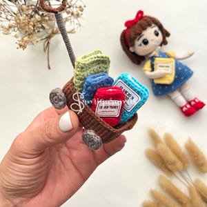 May include: A miniature crochet wagon filled with colorful books and a doll. The wagon is brown with gray wheels and a brown handle. The books are green, blue, and red, with labels. A doll with a red bow and a blue dress is nearby.