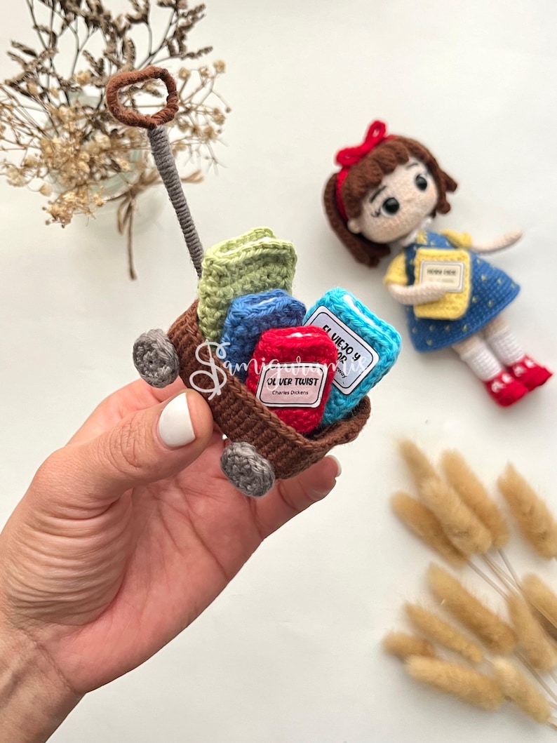May include: A miniature crochet wagon filled with colorful books and a doll. The wagon is brown with gray wheels and a brown handle. The books are green, blue, and red, with labels. A doll with a red bow and a blue dress is nearby.