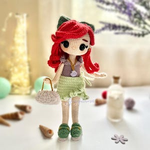 May include: A handmade crochet doll with red hair, a green bow, and a green skirt. The doll is wearing a purple top and green shoes. It is holding a small white purse with a gold handle. The doll is standing on a white surface.