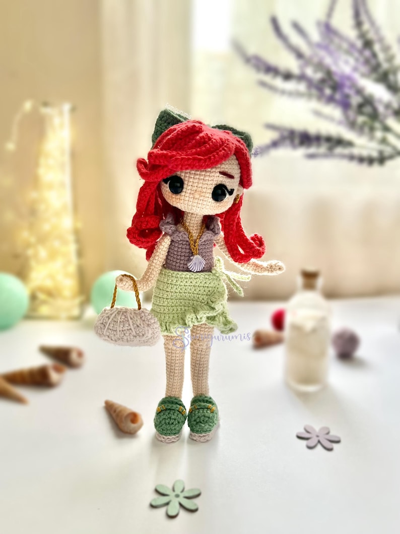 May include: A handmade crochet doll with red hair, a green bow, and a green skirt. The doll is wearing a purple top and green shoes. It is holding a small white purse with a gold handle. The doll is standing on a white surface.