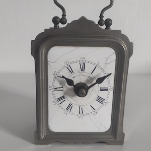 Puede incluir: Un reloj rectangular de estilo vintage con asa. La esfera del reloj presenta números romanos y manecillas negras. El marco y el asa son de color metálico oscuro. El reloj es probablemente un objeto decorativo.