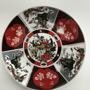 Puede incluir: Un plato de cerámica redondo con un medallón central que muestra dos pavos reales rodeados de flores coloridas. El borde exterior está dividido en seis secciones alternas, con patrones florales rojos y escenas en blanco y negro de carretas.