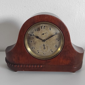 Puede incluir: Un reloj de sobremesa de madera vintage con la parte superior y la base curvadas. La esfera del reloj es dorada con números y manecillas negras. El reloj mide aproximadamente 25 cm de ancho y 18 cm de alto.