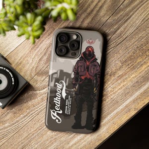 Op de afbeelding: Een grijze telefoonhoes met een Redhood-afbeelding. Het ontwerp bevat een figuur in een rode hoodie en het woord "Redhood" in een gestileerd lettertype. De hoes heeft een matte afwerking en een camera-uitsparing.