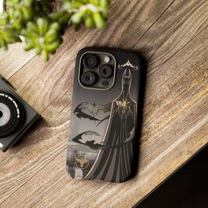May include: A black phone case with a graphic of Batman in a dark, stylized design. The case has a black background with gold accents and features a silhouette of Batman with bat wings in the background.