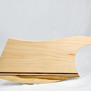 May include: A light-colored wooden cutting board with a unique curved design. The board features a handle with a hole for hanging and a decorative dark brown stripe. The board is approximately 18 inches long.