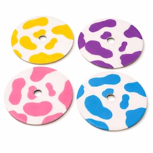 Puede incluir: Juego de cuatro posavasos redondos con base blanca y diseños de estampado de vaca de colores. Cada posavasos presenta un color diferente: amarillo, morado, rosa y azul. Tienen un agujero en el centro.