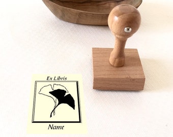 Può includere: Timbro in legno con un design a foglia di ginkgo e il testo "Ex Libris" e "Nome". Il timbro è su una base in legno e si trova accanto a una ciotola in legno.