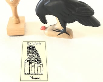 Può includere: Un timbro in legno con un design di corvo nero. Il corvo è appollaiato su un ramo e guarda in basso a una bacca rossa. Il timbro è su uno sfondo bianco. Il timbro è accanto a un timbro in legno con un design di gufo in bianco e nero. Il gufo è appollaiato su una pila di libri. Il timbro è su uno sfondo bianco. Il testo "Ex Libris" e "Nome" sono sul timbro.