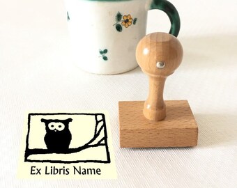 Può includere: Un timbro in legno con un'illustrazione in bianco e nero di un gufo su un ramo. Il timbro è montato su una base in legno e ha un manico rotondo in legno. Il timbro è etichettato "Ex Libris Name".