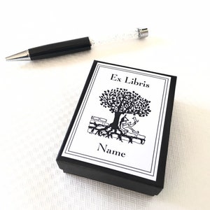 Könnte beinhalten: Ein schwarzer Stift mit einem transparenten, glitzergefüllten Schaft liegt neben einer schwarzen Box. Die Box hat ein weißes Etikett mit den Worten "Ex Libris" und einer Baumillustration. Das Wort "Name" steht darunter.