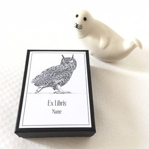 Puede incluir: Una caja negra con una etiqueta blanca que presenta una ilustraci&oacute;n en blanco y negro de un b&uacute;ho. La etiqueta dice "Ex Libris" y "Nombre".